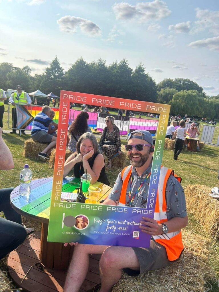 A sunny Pride event in a park, with people relaxing on hay bales and picnic tables behind a colourful Pride photo frame. In the foreground, two people smile for the camera beside a rainbow-coloured table, while a volunteer in a high-visibility vest holds the frame.